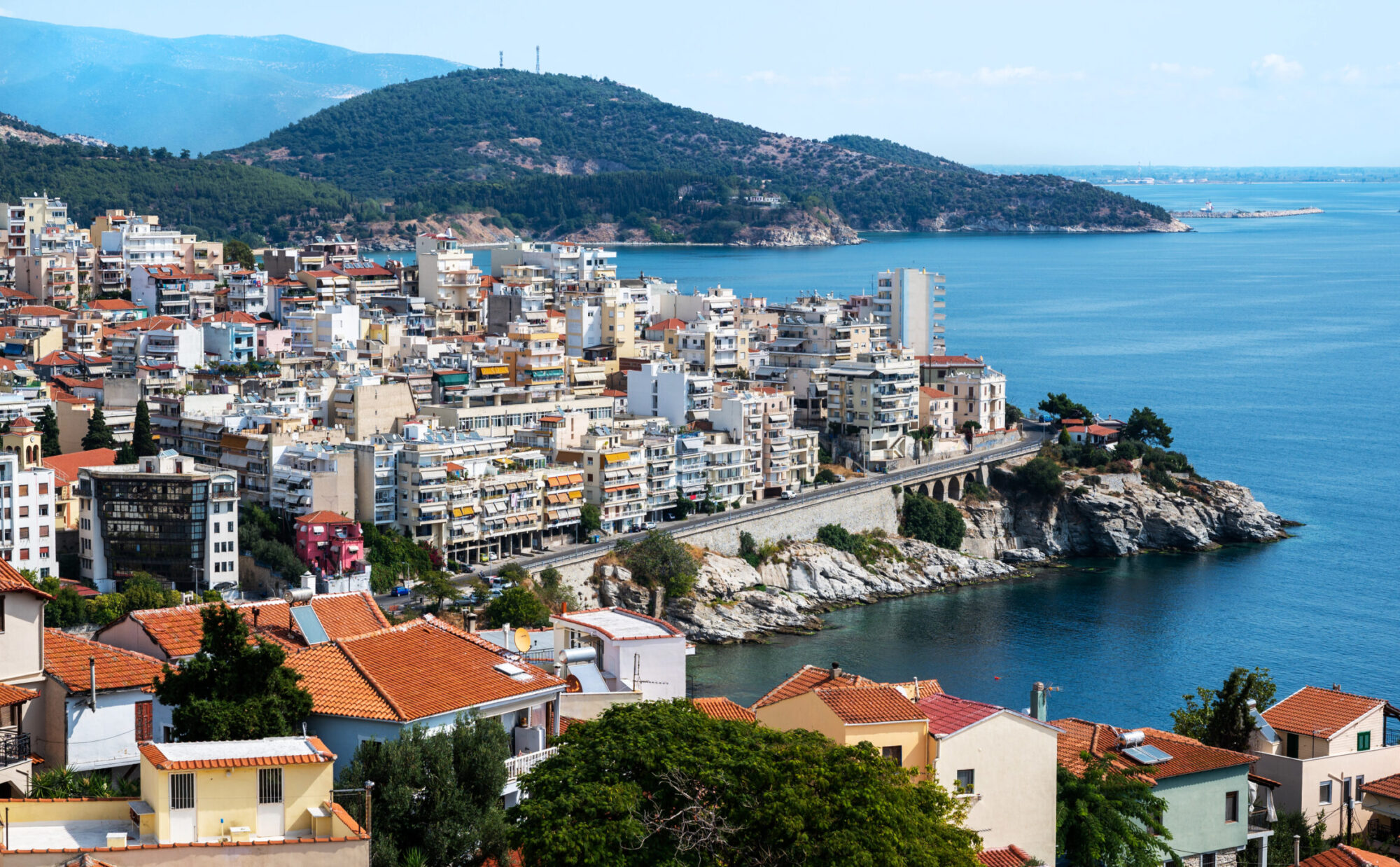 Multiple buildings located on the Aegean sea cost, road passing over the rocky coastline, green hills on the background in Kavala, Greece